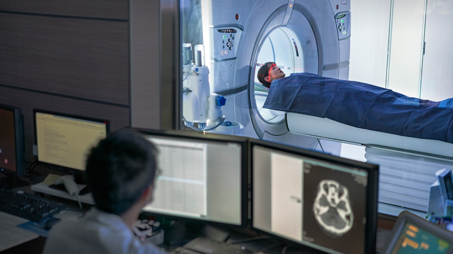 Older male healthcare professional looking at computer screens while younger female patient has MRI done